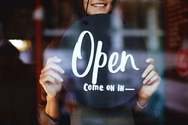 A store owner behind a glass door holding up a sign that says "Open, come on in"